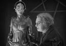 A white haired man stands in front of a female robot