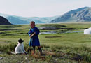 A girl stands in a field with a dog surrounded by mountains