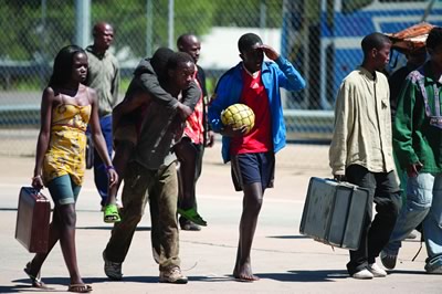 Four African children walk across concrete ground, one is being carried on another’s back.  Other people are walking behind and next to them some of whom are carrying cases.