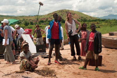 Three African children and a European woman stands on a film set, a sound boom and members of a film production crew are nearby.