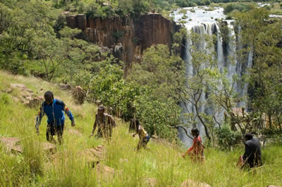 Wide shot of 5 African children walking up a hillside through long grass in front of a waterfall