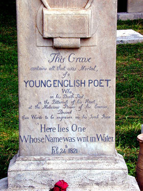 Close-up of a traditional headstone in a light sandy colour, a red flower placed in front. Keats' own words are clearly visible: 'Here lies one whose name was writ in water'