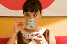 medium shot of actress with her dark hair tied back, eyebrows raised and looking left, drinking delicately from a teacup with one finger crooked