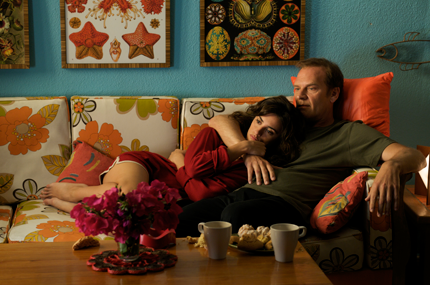 a couple on a sofa in a brightly coloured apartment have their arms around each other as they look thoughtfully off screen