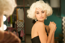 actress in platinum blonde bob wig and black evening dress stares at her reflection in the mirror, reminiscent of Marilyn Monroe