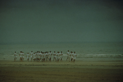 A group of young men in matching white running kits run in a line along a beach