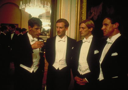 Four young men wearing tuxedos stand in a grand hall drinking champagne