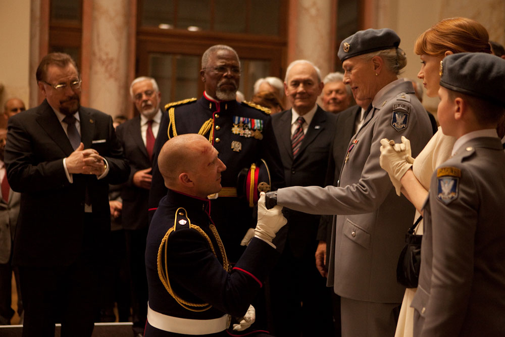 Mid-long shot of a group of people dressed in either suits or military regalia, around a central male figure who is kneeling and holding the hand of an older woman also in military dress uniform. A younger woman stands to her left; all characters are attendant on the central male.