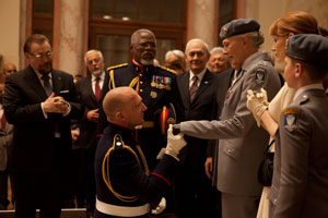 Mid-long shot of a group of people dressed in either suits or military regalia, around a central male figure who is kneeling and holding the hand of an older woman also in military dress uniform. A younger woman stands to her left; all characters are attendant on the central male