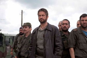 mid shot of eight men dressed in combat khakis looking off to the front left of the shot. The men are grouped around a central man who appears taller than the others; he ears a leather jacket over his uniform