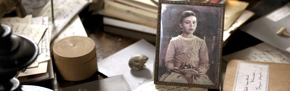 A desk covered in notebooks, hand-written papers and small collected specimens with a black and white photograph of a young girl centre-right.