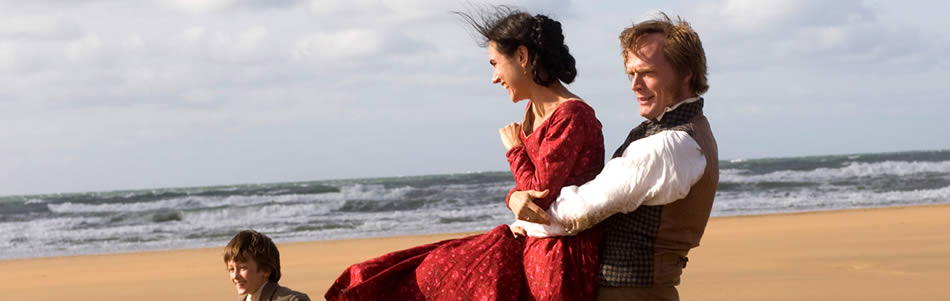 Charles Darwin lifting his wife Emma on the beach with the sea in the background. Their small son walks beside them.