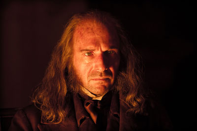 darkly lit mid-shot showing a man with long grey hair and period costume staring intently