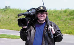 A man stands holding a video camera on his shoulder and a gun