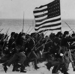 Still in black & white of U.S. soliders marching with guns and a U.S. flag on a beach