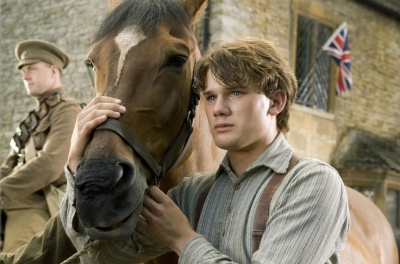 A boy stands holding a horse around its head with a soldier standing in the background