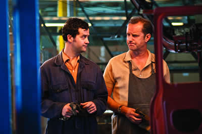 A man in a navy boiler suit stands on the left of the image and looks at a workmate in dungarees standing next to him.  The men are looking at each other with expressions suggesting they share an irritation or annoyance about something.  In the foreground to the right is part of a car door; to the left are painted blue iron girders.  There hanging flourescent lights behind them and blurred in the background are stacked large brown boxes.  
