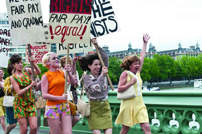 Five women at the forefront of a protest march across a bridge.  They look happy and are singing or chanting.  They are carrying banners reading: ‘RIGHTS. Fair pay!  Equal!  Fair pay and grading’.  The women are wearing 1960s style clothes such as miniskirts and hotpants. 