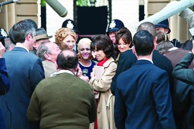 Rita stands smiling at the centre of the image wearing a beige trench coat and holding a folder. Three other women strikers accompany her and we can see all the women’s faces looking very happy.  Male reporters surround them and have their backs to the audience. Over their heads are two boom mikes and behind the women stand two uniformed police officers.