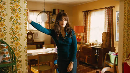 A woman in a blue dress leans against the wall in a 1970s style living room