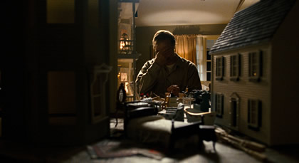 A man stands, covering his face with his hand, in a darkened room, with several large dolls houses