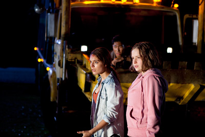 Two teenage girls stand in the foreground in front of a garbage truck, hesitating as they look at something out of shot. A teenage boy is in the bucket of the truck behind them, also looking out of shot.