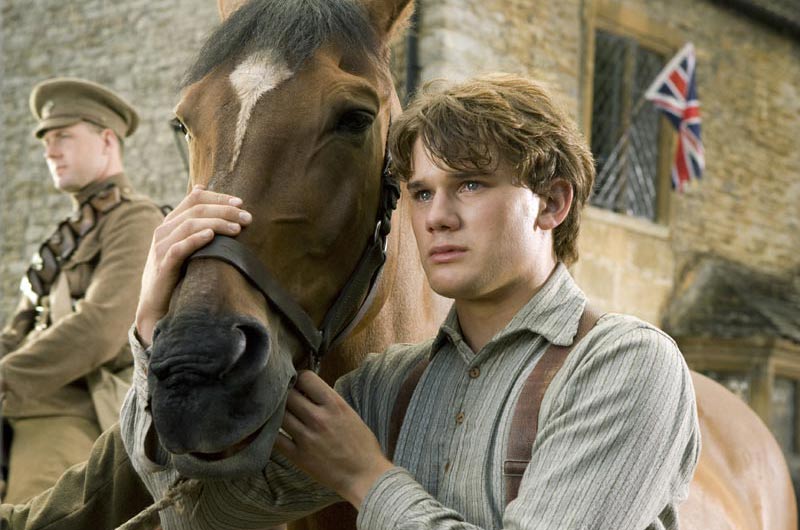 Head shot of young man wearing shirt and braces standing close to horse, holding the horse’s reins.