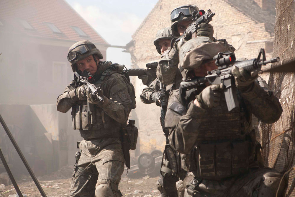 mid-long shot of four soldiers dressed in modern-style camouflage combat gear and helmets. Their automatic weapons are directed out of the shot to the right. Rubble and smoke, and domestic buildings, can be seen behind. 