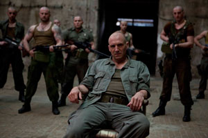 long shot shows armed men in khaki-coloured trousers and vests in the background; in the centre foreground, a seated man, also in khaki, with a commanding and stern expression looks out.