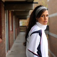 Young woman in foreground wearing loose vest top and gold necklace, scarf in her hair leaning against outside door of flat on an estate.  Next to her stands a young man wearing vest top and double chain gold necklace.  They are looking at something out of shot.