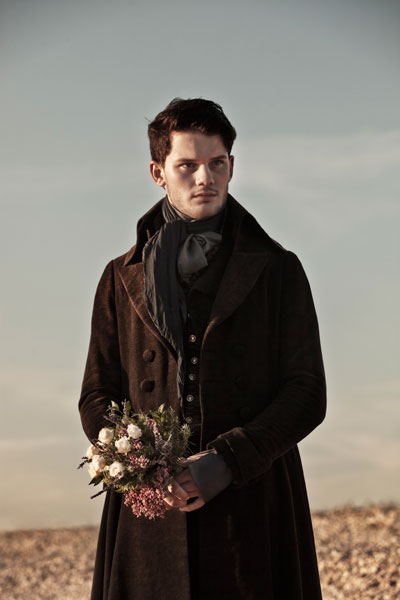 medium long shot of young male character in dark-coloured Victorian dress holding flowers