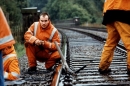 A man in an orange uniform kneels down on a train track with other men in the same uniform standing beside him