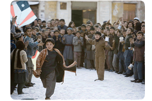 Still image from the film, 1 boy runs from another in a busy street, in which a large crowd watches them.