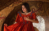 A young woman holds up a red gown against herself. She is standing in a cave-like dwelling with painted flowers decorating the walls