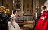 Five people stand in a smartly decorated room. There are two guards wearing shiny gold helmets that cover their faces. They are stood either side of a young woman dressed in a white gown. Opposite them is a man in period dress and a fancy dress 'antennae' hat. Next to him is a woman with her hands on her hips, wearing a scarlet dress with a white peacock tail attached to the back