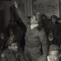 A black pilot holding a knife standing in a room surrounded by other pilots