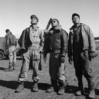 Three African American airmen stand together looking skyward. Another airman stands beside a truck in the background