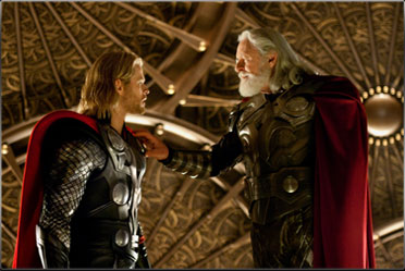 An older bearded man and younger clean shaven man wearing armour and cloaks. He has long blond hair.